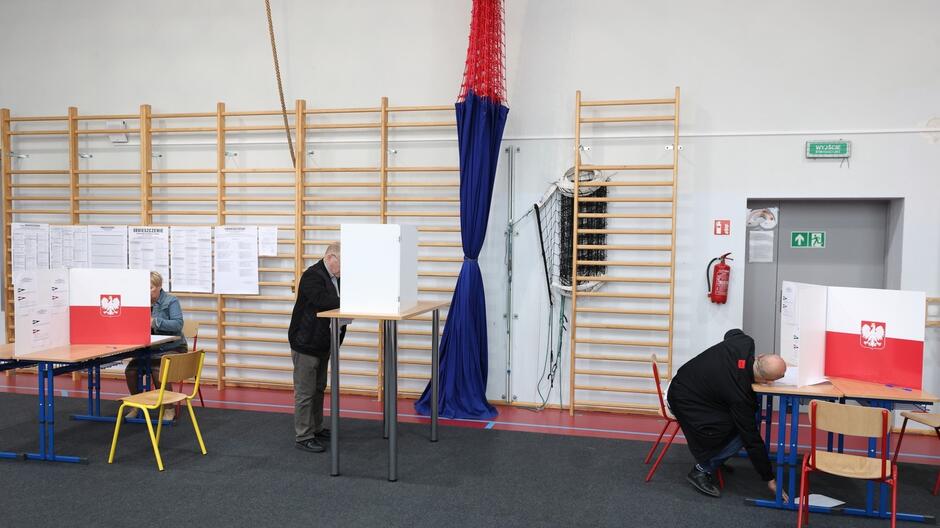 Na zdjęciu widzimy wnętrze hali sportowej, która została zaadaptowana na miejsce do głosowania. Na ścianie zawieszone są drabinki gimnastyczne, a między nimi rozwieszona jest duża, niebiesko-czerwona siatka. W centrum zdjęcia widoczne są trzy kabiny do głosowania, każda z nich ma na przodzie umieszczone duże białe tablice z polskim godłem – białym orłem na czerwonym tle. Po lewej stronie zdjęcia siedzi kobieta przy stole obok niebieskiego krzesełka, wydaje się, że pełni funkcję członka komisji wyborczej. Po prawej stronie, mężczyzna w czarnej kurtce zagląda do urny wyborczej, sprawdzając prawidłowość jej zamknięcia. W tle, na ścianie, zawieszone są liczne listy i obwieszczenia prawdopodobnie związane z wyborami. Na podłodze, pod kabinkami wyborczymi, leżą różnokolorowe linie, które zwykle wyznaczają obszary na sali gimnastycznej. Pomieszczenie jest dość jasne i przestronne
