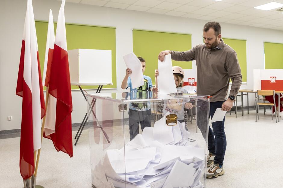 Na zdjęciu widać wnętrze jasnego pomieszczenia, prawdopodobnie szkoły lub innego budynku publicznego, gdzie odbywa się proces głosowania. Mężczyzna wkłada swój głos do przezroczystej urny wyborczej. Obok niego stoją dwoje małych dzieci, które również trzymają w rękach karty do głosowania, być może pomagając dorosłym. W tle widoczne są polskie flagi oraz kabiny do głosowania z emblematem Polski na białych parawanach. Cała scena wydaje się być momentem zaangażowania obywatelskiego, z udziałem różnych pokoleń. Atmosfera jest spokojna i oficjalna.