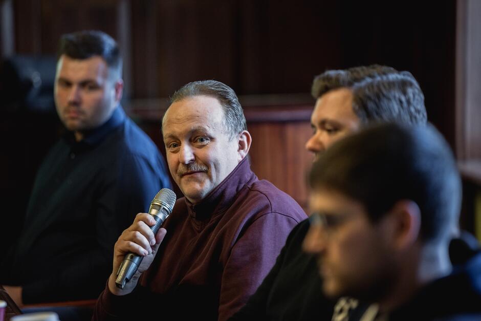 na zdjęciu mężczyzna w średnim wieku, w ciemnym swetrze, mówi do mikrofonu, siedzi, obok niego siedzi trzech innych mężczyzn