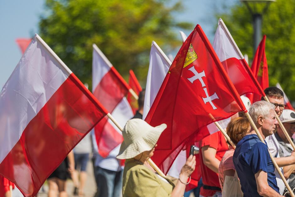 Na zdjęciu widać grupę ludzi uczestniczących w wydarzeniu na otwartym powietrzu, gdzie trzymają one różne flagi. Są to flagi białe oraz czerwone, z których niektóre mają dodatkowe symbole, w tym herb z białymi krzyżami. Tło zdjęcia zdobi jasne, letnie niebo oraz zielone drzewa, co sugeruje, że wydarzenie odbywa się w ciepły, słoneczny dzień. Uczestnicy wydają się być skoncentrowani na wydarzeniu, obserwując coś poza kadrem zdjęcia. Atmosfera wydaje się spokojna i uroczysta.