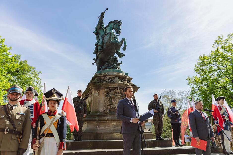 na zdjęciu mężczyzna w garniturze mówi do mikrofonu obok niego stoją żołnierze i rekonstruktorzy w historycznych strojach wojskowych, w tle pomnik króla na koniu