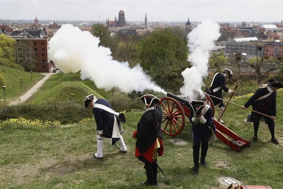 Na zdjęciu widać grupę osób ubranych w historyczne mundury, które obsługują działko z czasów napoleońskich. Scena rozgrywa się na zielonym wzgórzu, z którego roztacza się widok na miasto w tle. Osoby są ubrane w białe spodnie, granatowe kurtki z czerwonymi detalami i czarne trójgraniaste kapelusze, co jest charakterystyczne dla wojskowego stroju z epoki napoleońskiej. Działko właśnie zostało wystrzelone, co widać po gęstym białym dymie wypływającym z lufy. To historyczna rekonstrukcja lub pokaz, który ma na celu przedstawienie metody działania i obsługi artylerii z tamtych czasów.