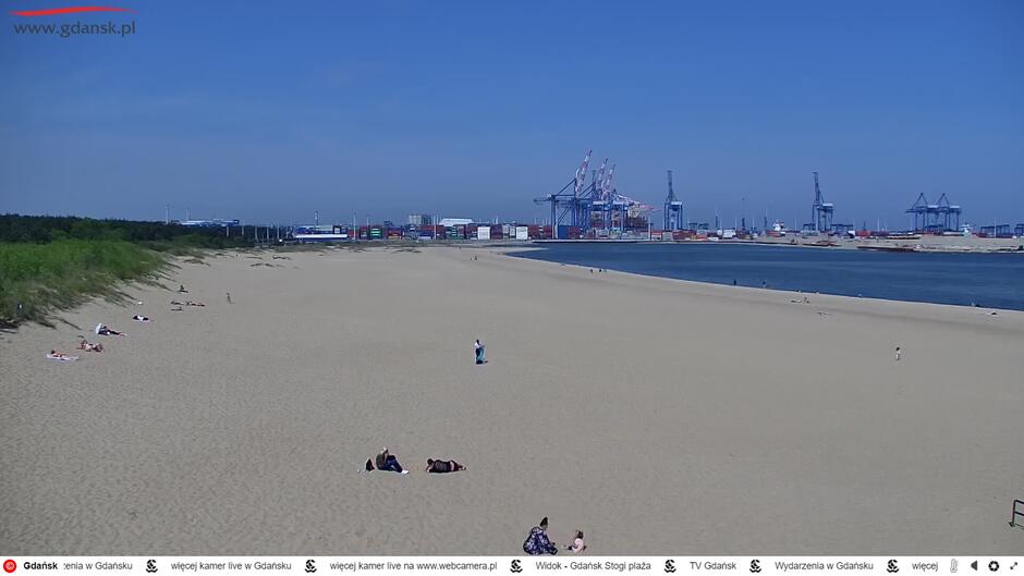 Zrzut ekranu z kamery miejskiej. Widok na plażę, na której leży kilka osób. W tle dźwigi portowe.