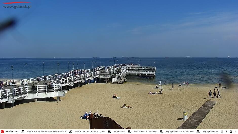 Zrzut ekranu z kamery miejskiej. Widok na plażę, na której leży kilkanaście osób oraz białe, drewniane molo.