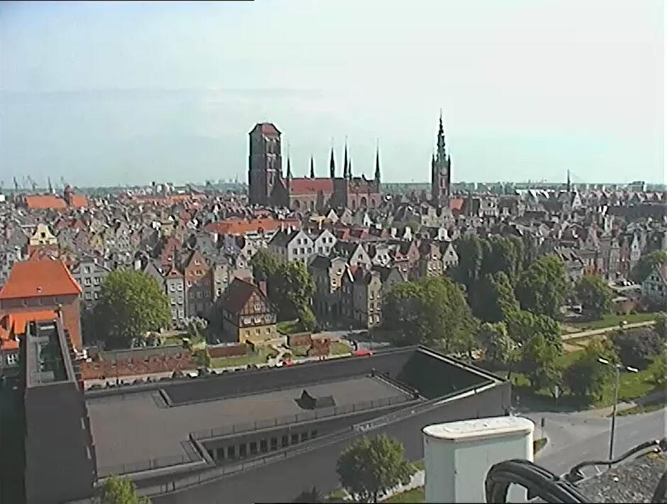 Zrzut ekranu z kamery miejskiej. Widok na czarną bryłę teatru na pierwszym planie. W tle stare kamienice oraz wieże - kościoła i ratusza.