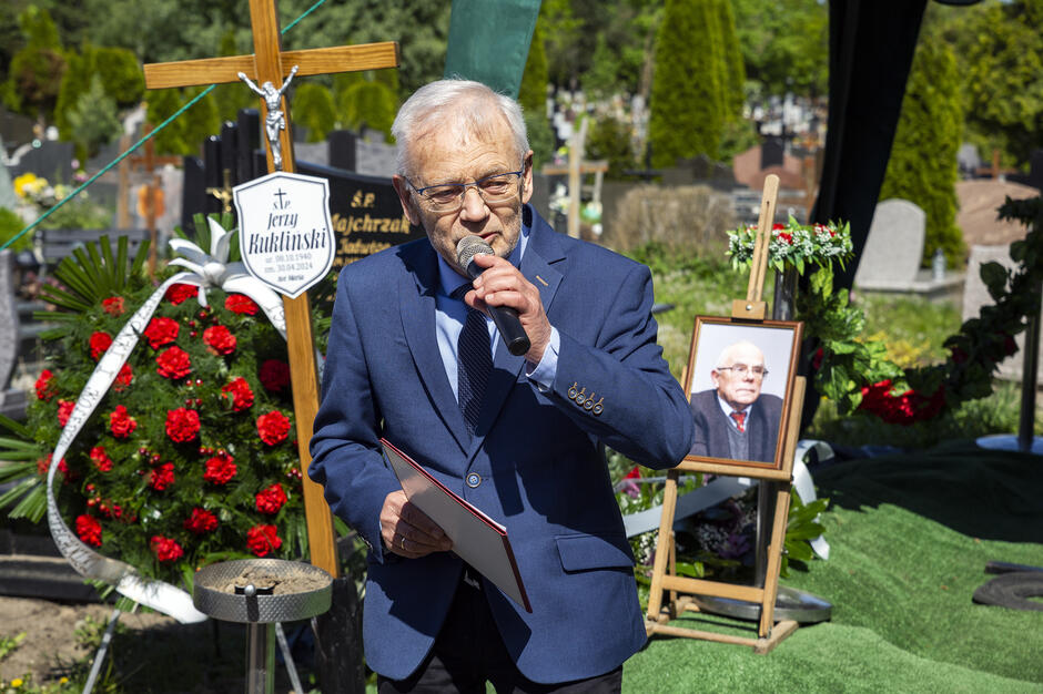 Mężczyzna z mikrofonem przemawia na pogrzebie. W tle krzyż i zdjęcie zmarłego na stojaku. 