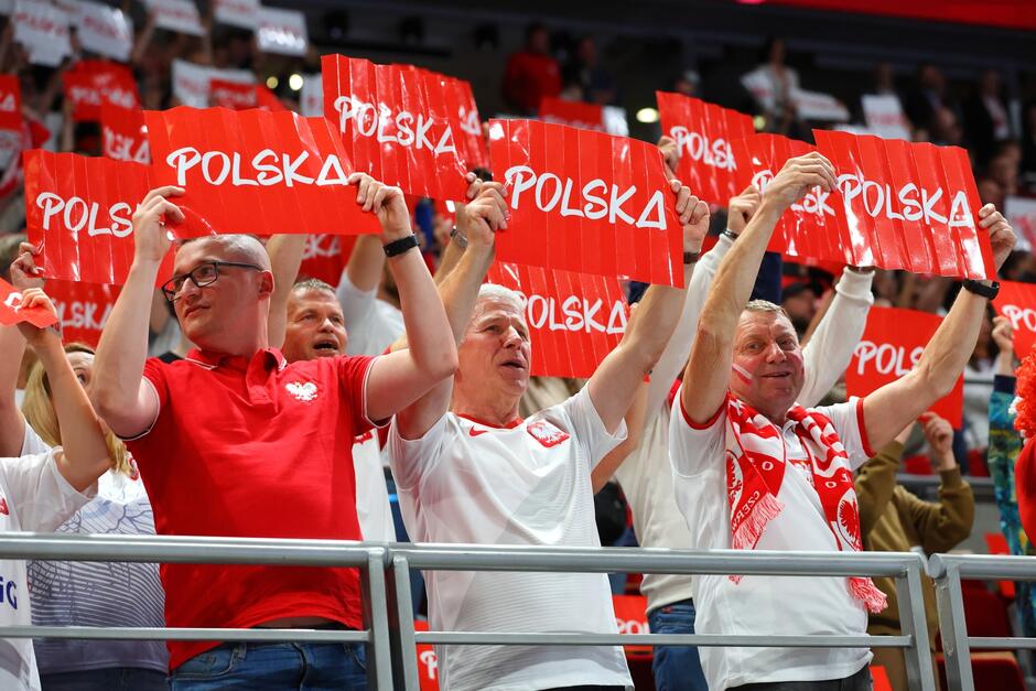 Na zdjęciu widać grupę polskich kibiców, którzy entuzjastycznie dopingują, trzymając w górze czerwone kartki z napisem POLSKA . Są ubrani w narodowe barwy – czerwone koszulki, niektórzy mają także białe elementy, jak koszulki i szaliki z orłem, symbol narodowy Polski. 