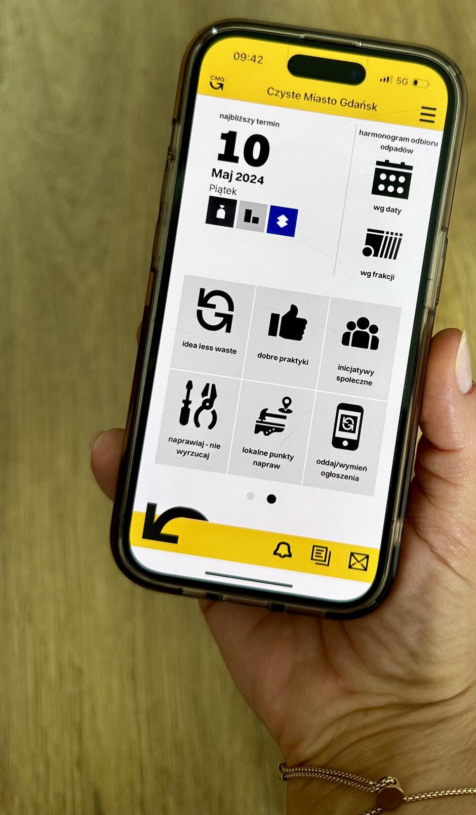 Zdjęcie przedstawia aplikację Czyste Miasto Gdańsk  wyświetlaną na smartfonie trzymanym w dłoni osoby. Ekran główny aplikacji jest zaprojektowany w estetyce z dominującymi żółtymi i czarnymi kolorami, co czyni interfejs wyraźnym i czytelnym.

Na ekranie wyświetlone są różne ikony funkcji aplikacji:

Najbliższy termin odnośnie daty ( 10 Maj 2024, Piątek ) z ikonami dotyczącymi śmieci, które mają zostać odebrane tego dnia.
Harmonogram odbioru odpadów.
Ikonę  idea less waste  sugerującą funkcje związane z minimalizacją odpadów.
Ikonę  dobre praktyki , która prawdopodobnie prowadzi do porad na temat zrównoważonego życia.
Sekcję  inijcatywy społeczne  z ikoną grupy ludzi, wskazującą na funkcje społecznościowe aplikacji.
Opcję  naprawiaj - nie wyrzucaj  z narzędziami, sugerującą funkcje pomagające znaleźć lokalne punkty napraw.
Ikonę  oddaj/wymień ogłoszenia , która najprawdopodobniej umożliwia użytkownikom dodawanie ogłoszeń o wymianie lub oddaniu przedmiotów.
Aplikacja jest przykładem narzędzia cyfrowego, które ma na celu wspieranie działań na rzecz zrównoważonego rozwoju i minimalizacji odpadów poprzez praktyczne rozwiązania i społeczność lokalną.