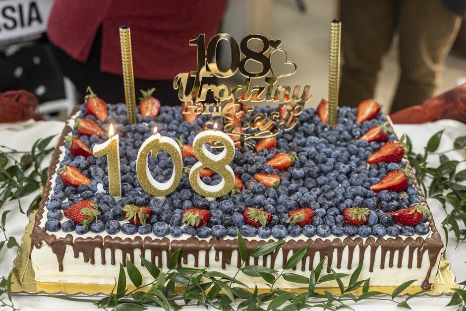 Zdjęcie przedstawia uroczyście przygotowany tort urodzinowy z okazji 108. urodzin, obficie pokryty borówkami i truskawkami. Na torcie umieszczone są złote ozdoby z napisami „108” oraz „108 trochę trwało, ale się doczekało”
