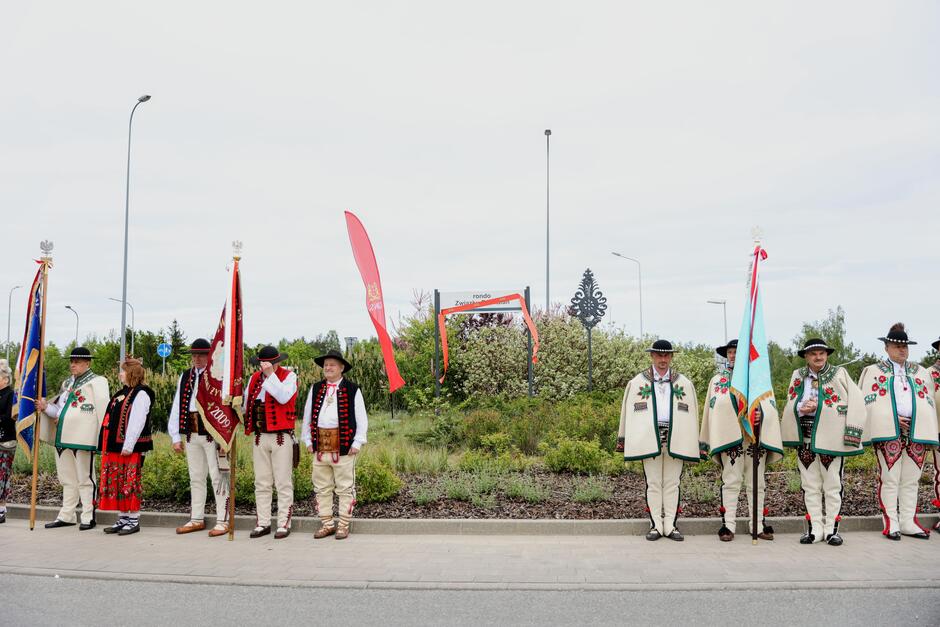 Na zdjęciu grupa mężczyzn w tradycyjnych, regionalnych strojach stoi obok siebie, trzymając sztandary i flagi. Tło zdjęcia to nowoczesny krajobraz miejski z drogowymi znakami i niewielkimi drzewami, co tworzy kontrast między tradycją a współczesnością