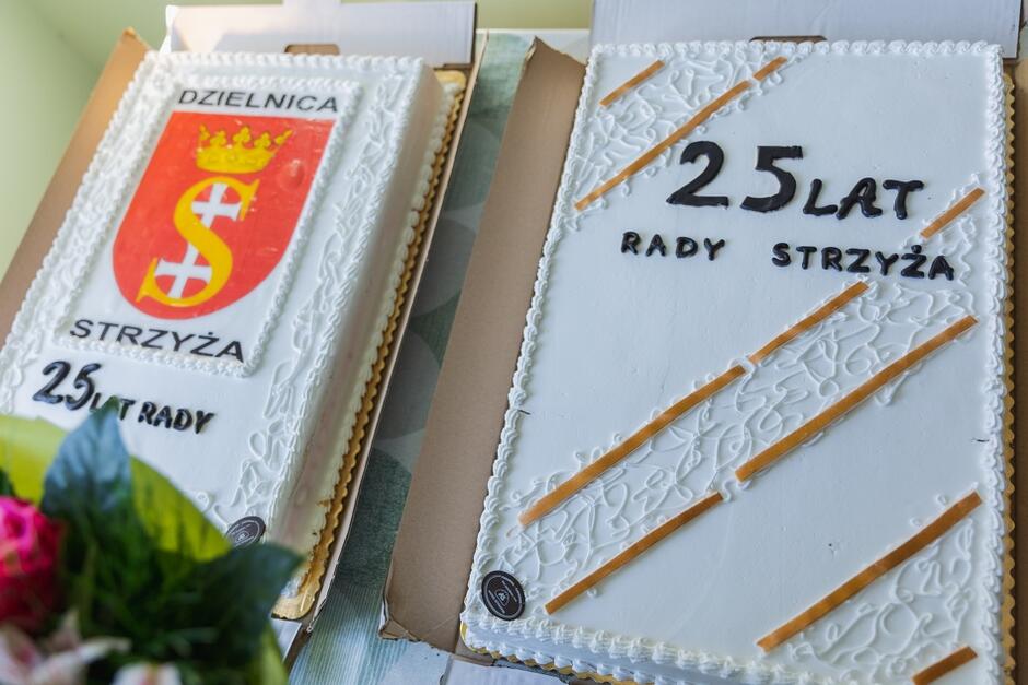 Na zdjęciu są dwa torty z okazji 25-lecia Rady Dzielnicy Strzyża. Jeden tort jest ozdobiony herbem dzielnicy, a drugi napisem „25 lat Rady Strzyża” oraz dekoracyjnymi, jasnobrązowymi pasami






