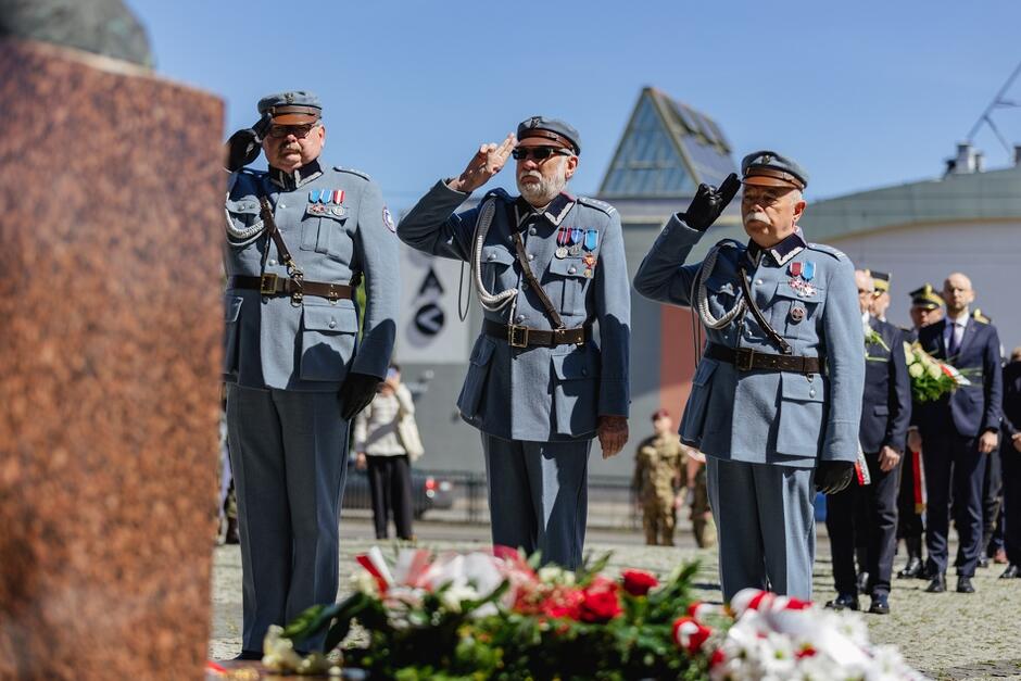 Na zdjęciu widać trzech mężczyzn w szarych wojskowych mundurach, którzy oddają honory, salutując podczas ceremonii. Są odznaczeni różnymi medalami i odznakami, a w tle widoczne są kwiaty oraz współczesne budynki, co sugeruje, że uroczystość odbywa się w miejskiej przestrzeni.