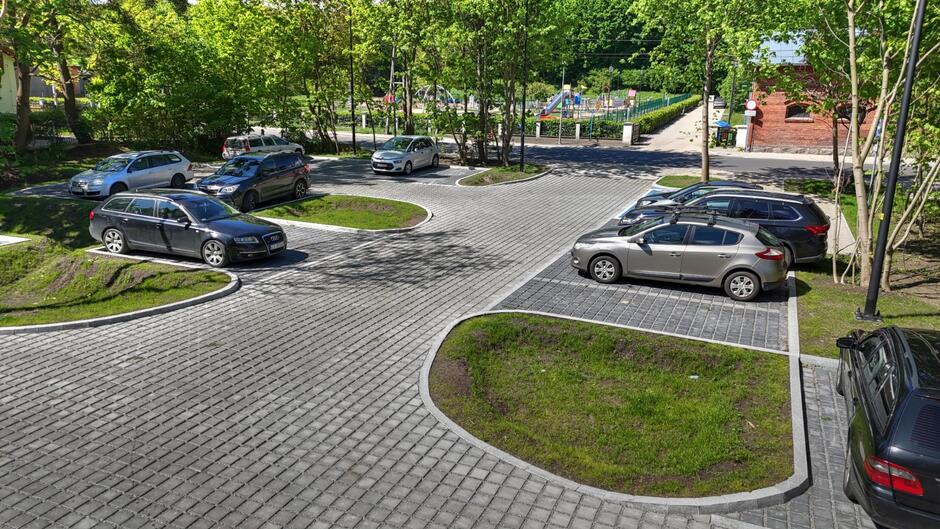 na zdjęciu fragment parkingu, widać nawierzchnię z kostki, kilka zaparkowanych samochodów osobowych i niewielkie zielone trawniki