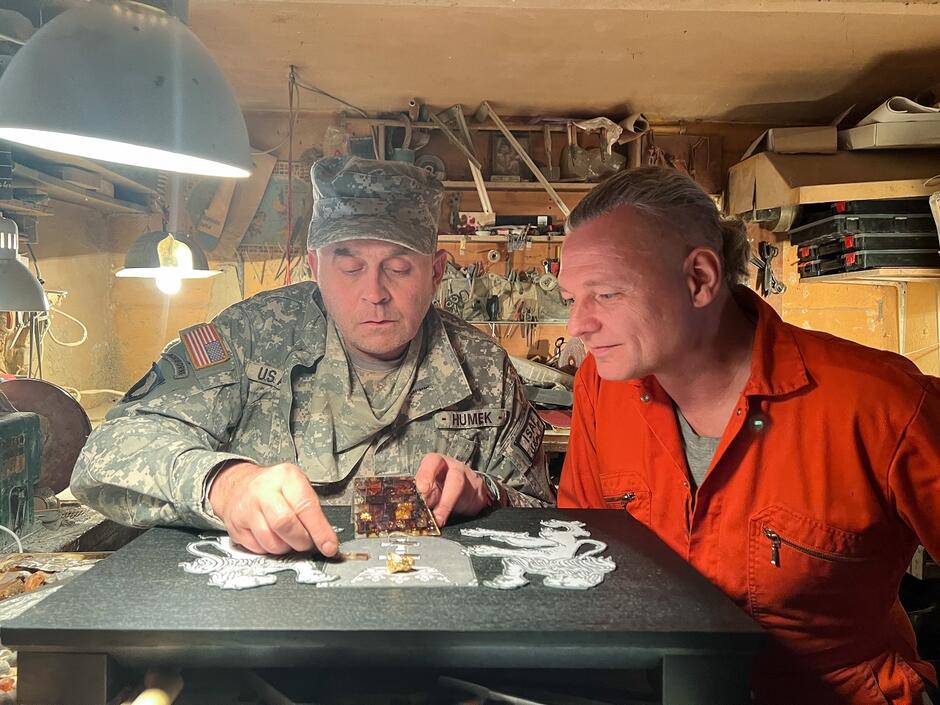 Na zdjęciu znajdują się dwaj mężczyźni pracujący w warsztacie. Jeden z nich nosi mundur wojskowy, a drugi ubrany jest w pomarańczowy kombinezon roboczy; obaj skupieni są na wspólnej pracy nad ozdobnym projektem na stole.