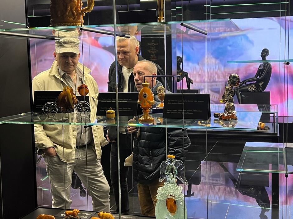  Na zdjęciu widzimy trzech mężczyzn oglądających wystawę w muzeum, skupiających się na eksponatach wykonanych z bursztynu. Ekspozycja zawiera różnorodne bursztynowe przedmioty, umieszczone w szklanych gablotach, a tłem jest kolorowa, rozmyta projekcja.