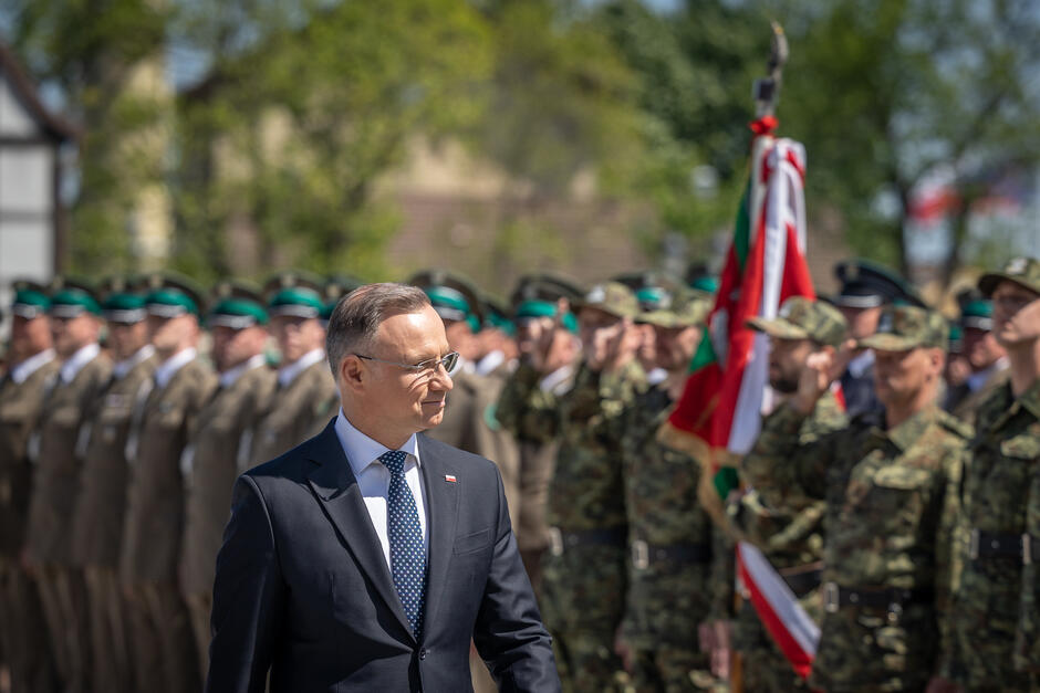 na zdjęciu prezydent polski w ciemnej marynarce i w okularach, ma głowę skierowaną w kierunku stojących w kilku rzędach strażników granicznych ubranych w galowe mundury