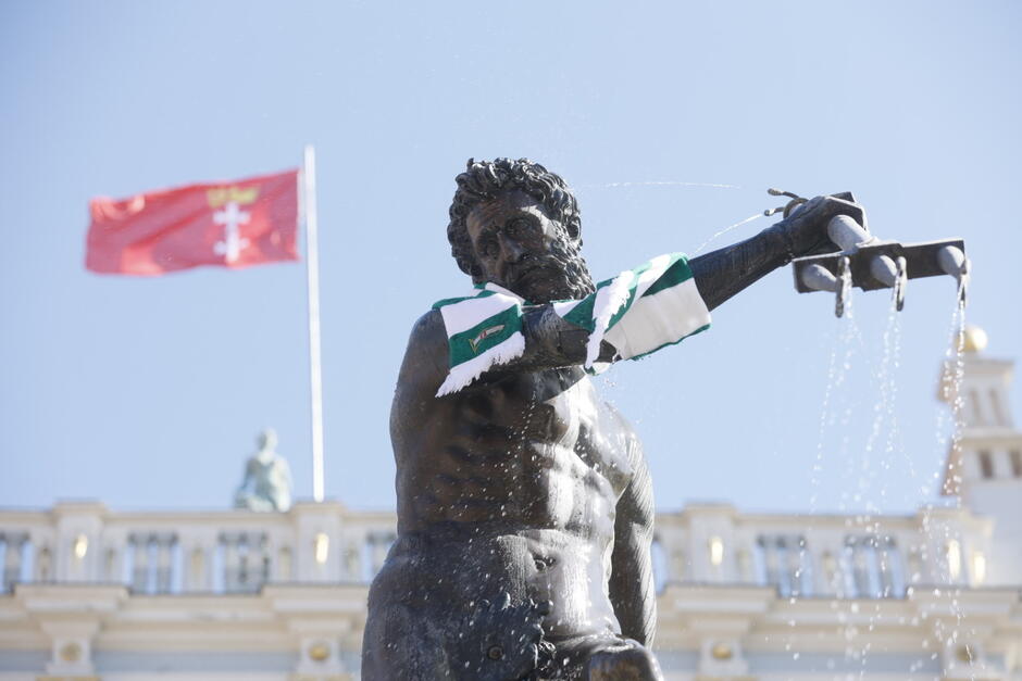Gdański Neptun w biało-zielonym szaliku. W tle flaga z herbem Gdańska. 