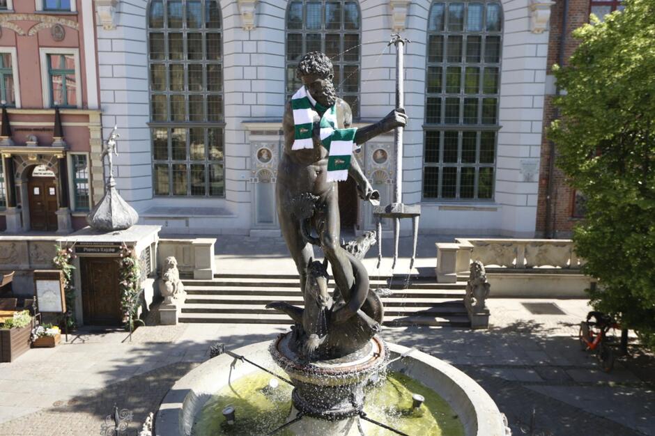 Figura Neptuna na fontannie z założonym na szyi biało zielonym szalikiem. W tle renesansowy budynek Dworu Artusa.