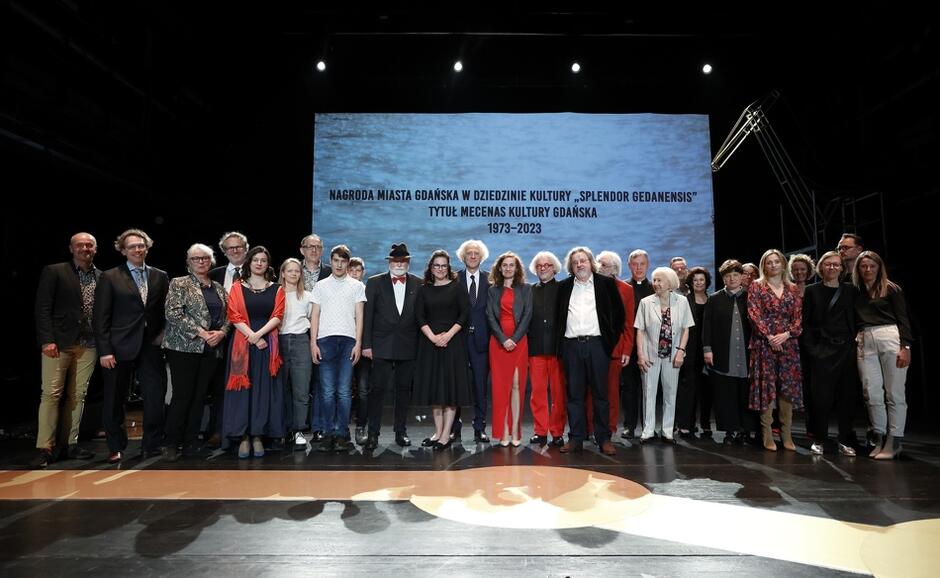 Na zdjęciu znajduje się grupa osób, które pozują na scenie podczas uroczystości wręczenia Nagrody Miasta Gdańska w Dziedzinie Kultury "Splendor Gedanensis". W tle widoczny jest ekran z napisem "Nagroda Miasta Gdańska w Dziedzinie Kultury 'Splendor Gedanensis' Tytuł Mecenasa Kultury Gdańska 1973-2023"