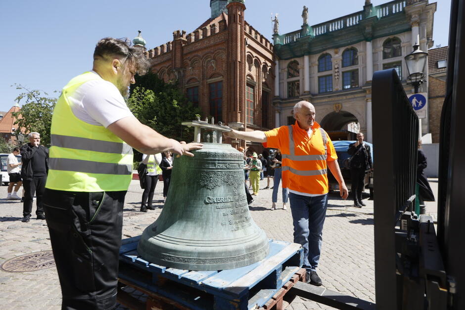 Zabytkowy dzwon stoi na drewnianej palecie, którą trzyma wózek widłowy. Obok dzwonu stoi dwóch mężczyzn w roboczych odblaskowych kamizelkach. W tle zabytkowe budynki Gdańska.