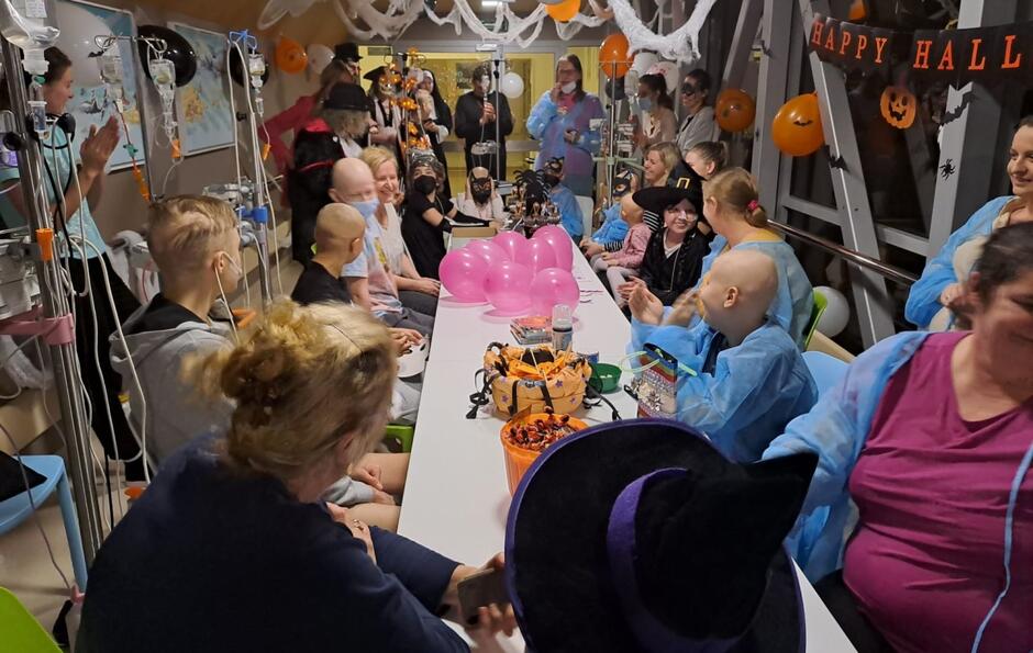 Na zdjęciu widać grupę dzieci i dorosłych, zebranych wokół długiego stołu w sali szpitalnej, podczas obchodów Halloween. Uczestnicy są przebrani w kostiumy, a na stole znajdują się różne dekoracje halloweenowe, takie jak różowe balony, cukierki i ozdoby, a ściany są ozdobione pajęczynami oraz napisami "Happy Halloween".
