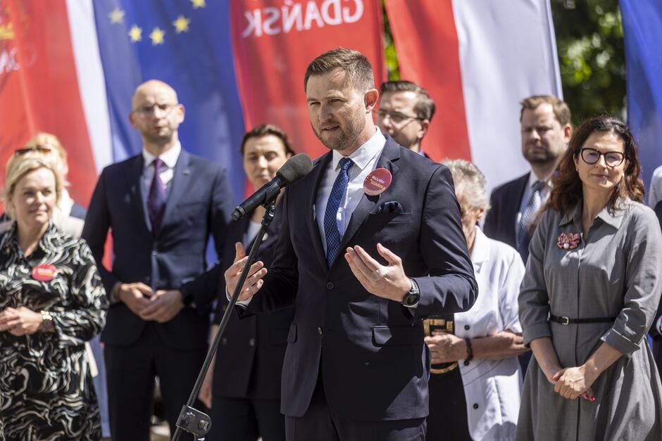 Na zdjęciu widzimy grupę ludzi, którzy biorą udział w wydarzeniu na świeżym powietrzu. W centralnej części znajduje się mężczyzna w garniturze, z przypiętym identyfikatorem, przemawiający do mikrofonu. W tle stoją inni uczestnicy, zarówno mężczyźni, jak i kobiety, również elegancko ubrani. W tle widoczne są flagi Unii Europejskiej oraz polskie flagi z napisem "GDAŃSK". Wydarzenie prawdopodobnie ma charakter oficjalny, być może jest to konferencja prasowa, wiec wyborczy lub inna uroczystość publiczna.