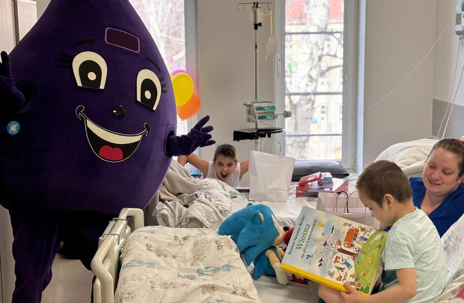 Na zdjęciu znajduje się chłopiec w szpitalnym łóżku, który czyta książkę, obok niego siedzi uśmiechnięta kobieta, prawdopodobnie jego mama. Obok łóżka stoi maskotka w kształcie fioletowej kropli krwi z szerokim uśmiechem, a w tle inny chłopiec robi zabawną minę.