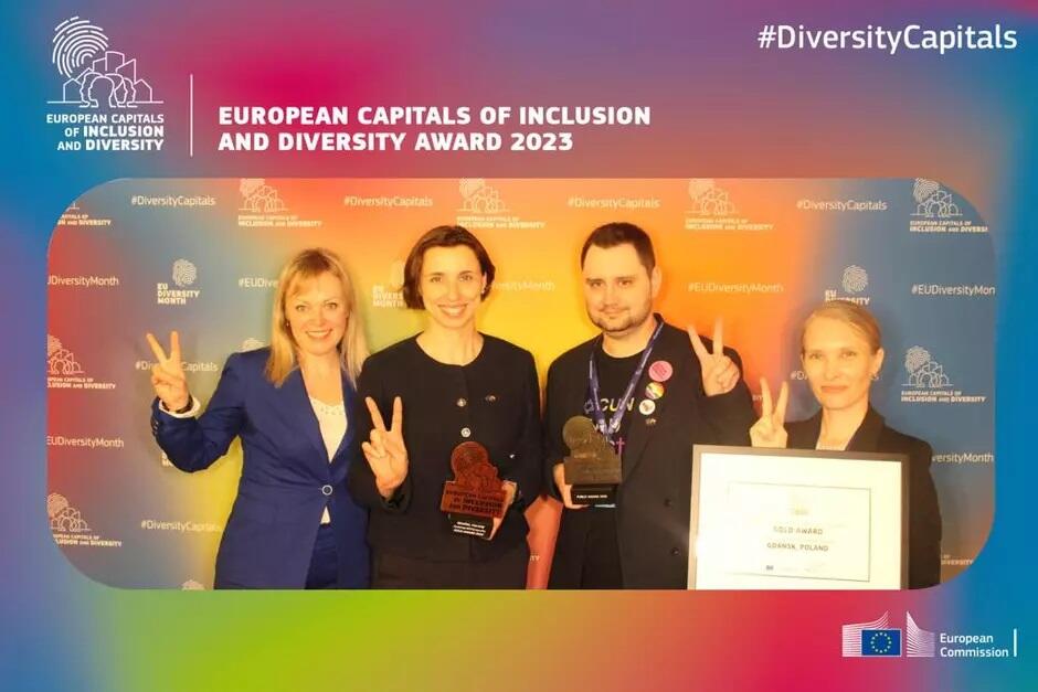 Grafika przedstawiająca nagrodę Europejskie Stolice Włączenia i Różnorodności . Na grafice umieszczono zdjęcie, na którym stoją obok siebie cztery uśmiechnięte osoby - trzy kobiety i mężczyzna, które pokazują palcami znak zwycięstwa.