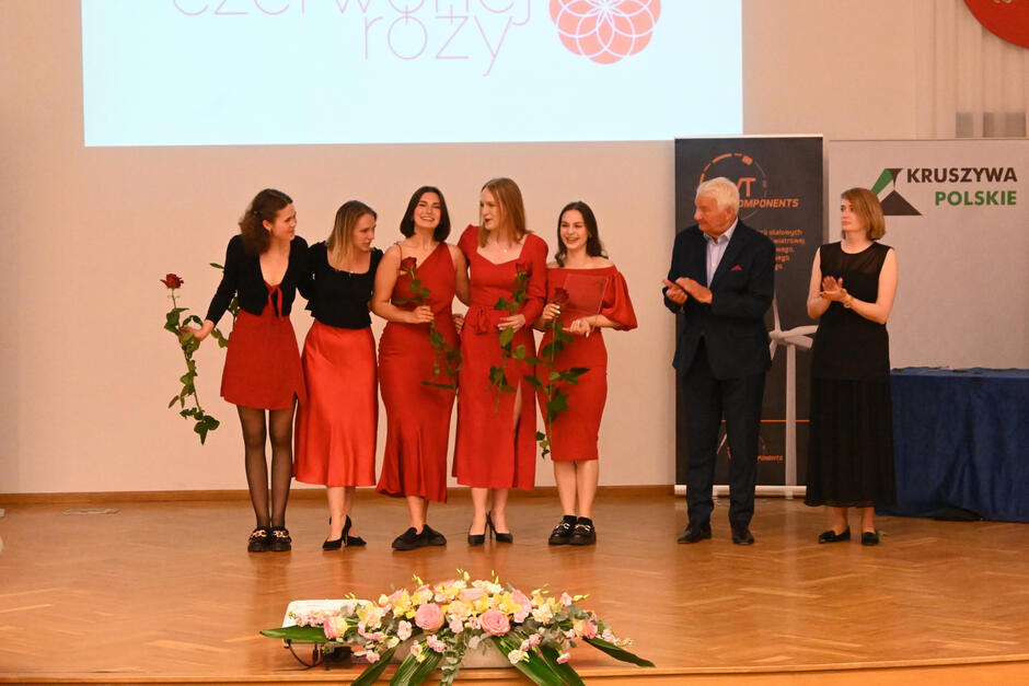 Pięć młodych kobiet stoi obok siebie na scenie, śmiejąc się. W rękach trzymają kwiaty, czerwone róże. jedna z nich trzyma nagrodę. Obok na scenie biją im brawo kobieta i mężczyzna.