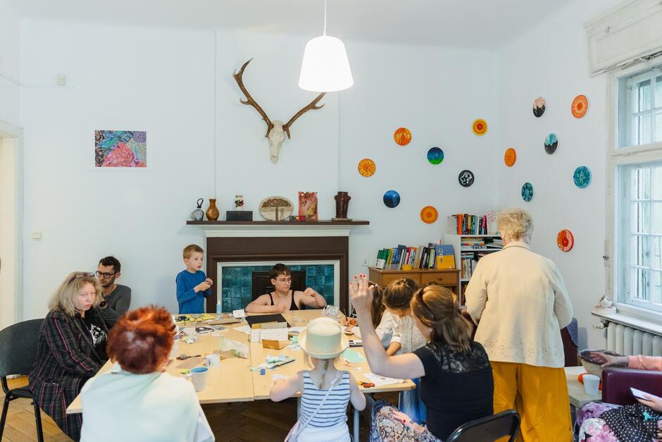 na zdjęciu stół przy którym siedzi kilkanaście osób, zarówno starsze kobiety, jak i osoby w średnim wieku oraz dzieci, niektóre coś wyklejają, ktoś coś czyta 