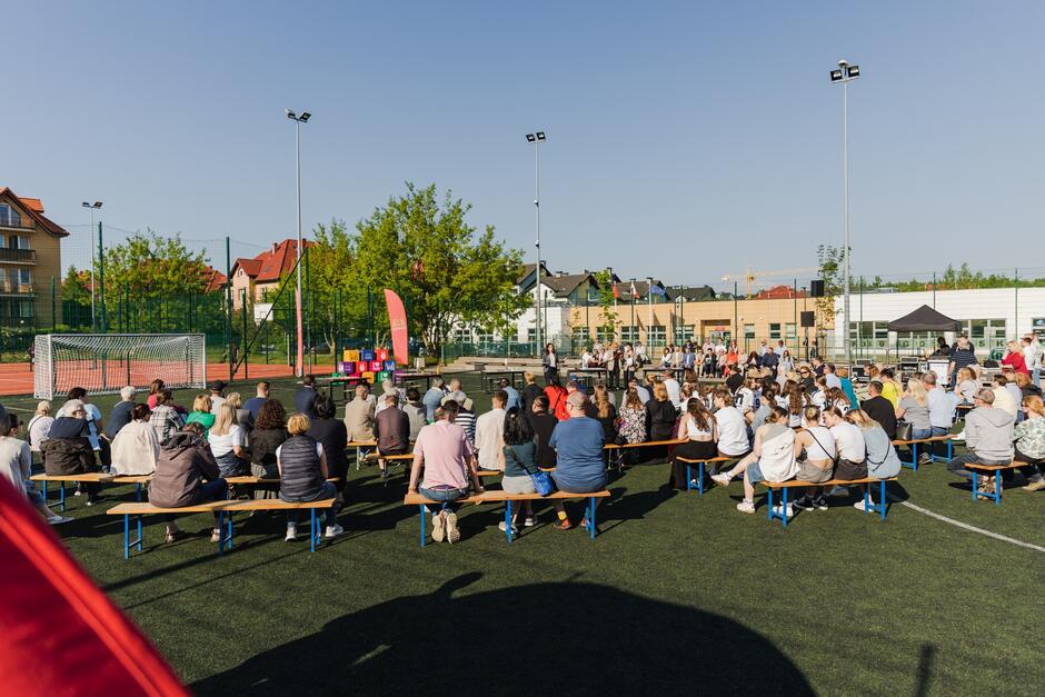 na zdjęciu boisko szkolne z zieloną murawą, widać po lewej bramkę, na boisku widać kilkadziesiąt osób siedzących na ustawionych specjalnie ławkach