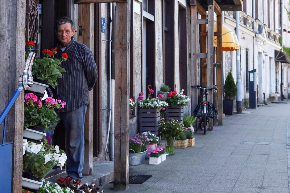 mężczyzna opiera się o framugę drzwi niewielkiej kwiaciarni, przed nią donice z kwiatami, w tle uliczka