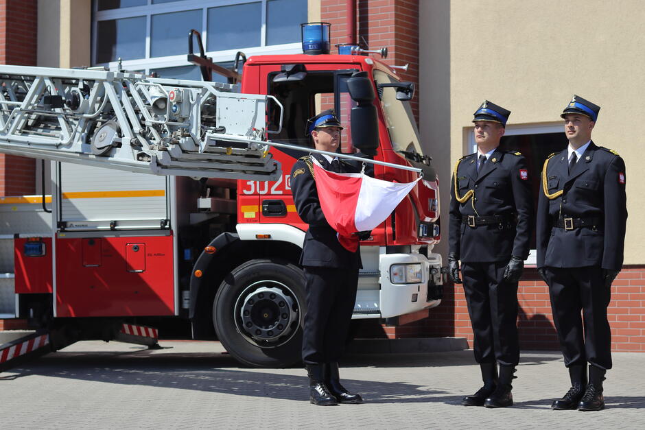 Na zdjęciu widzimy trzech strażaków w galowych mundurach stojących przed wozem strażackim z wysuniętą drabiną, z których jeden trzyma złożoną flagę Polski. W tle znajduje się budynek strażnicy, co sugeruje, że odbywa się uroczystość lub ceremonia związana z Dniem Strażaka