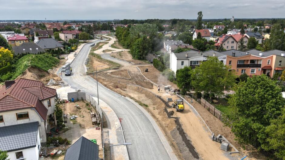 zdjęcie z drona, po lewej widać asfaltową jezdnię, po prawej budowaną jezdnię, widać na niej sprzęt budowlany, w tym koparki, przy ulicach stoją budynki jednorodzinne