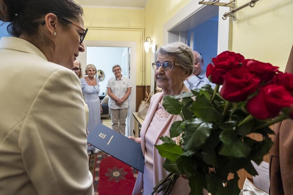solenizantka z bukietem kwiatów i dyplomem, prezydent obok, mówi coś do niej, w tle rodzina