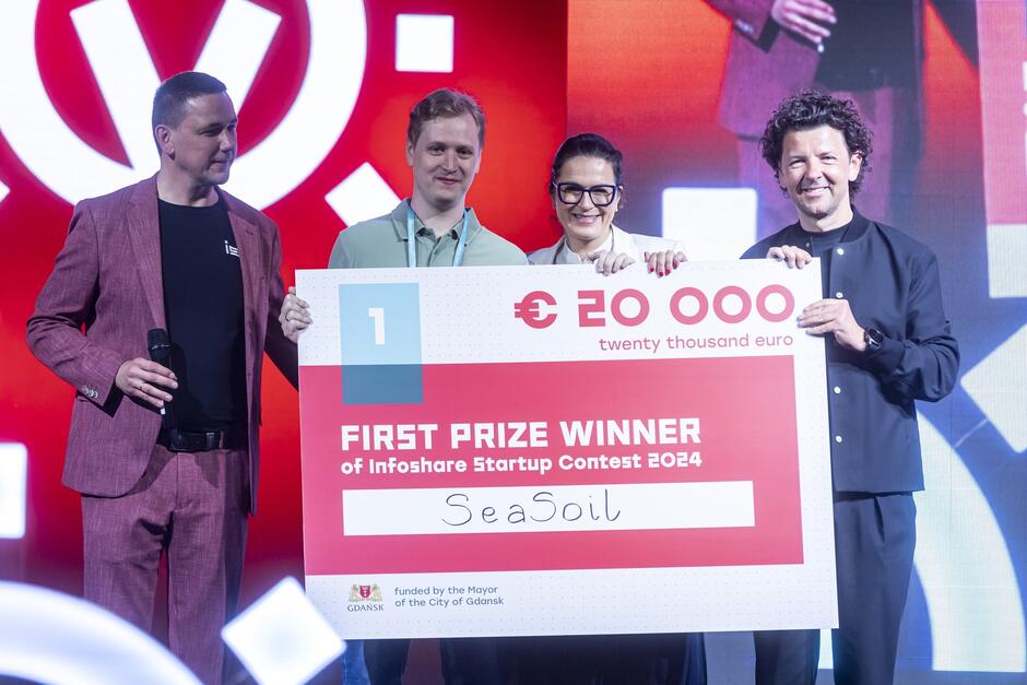 Na zdjęciu znajduje się cztery osoby na scenie, trzymające duży czek symbolizujący nagrodę za pierwsze miejsce w konkursie startupowym Infoshare Startup Contest 2024. Czek opiewa na kwotę 20 000 euro. Na czeku widnieje napis FIRST PRIZE WINNER of Infoshare Startup Contest 2024  oraz nazwa zwycięskiego startupu  SeaSoil . Osoba po lewej stronie trzyma mikrofon i jest ubrana w ciemny t-shirt i czerwono-brązową marynarkę. Osoba obok trzymająca czek jest ubrana w jasną koszulkę polo. Obok niej stoi kobieta w białej marynarce i ciemnych okularach. Osoba po prawej stronie ubrana jest w czarną marynarkę i trzyma czek z drugiej strony. Tło sceny jest ozdobione dużym logo konkursu oraz elementami graficznymi w czerwonych i białych barwach. Wszyscy na zdjęciu wyglądają na zadowolonych i dumni z wygranej.