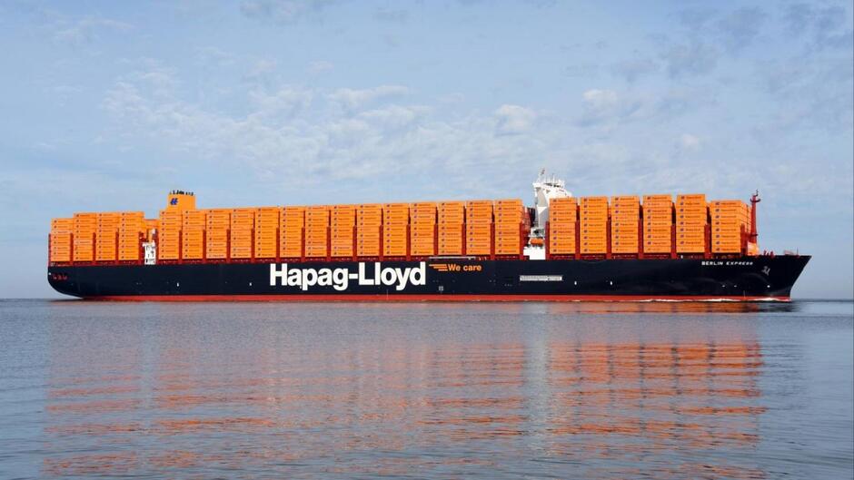 Na zdjęciu znajduje się ogromny kontenerowiec należący do firmy Hapag-Lloyd, pływający po spokojnych wodach morza. Statek jest załadowany dużą ilością jasnopomarańczowych kontenerów, które są ułożone w równych rzędach na pokładzie. Na burcie statku widnieje duży, biały napis Hapag-Lloyd  oraz hasło  We care , a na rufie statku można dostrzec nazwę  BERLIN EXPRESS . Statek jest pomalowany na ciemnoniebieski kolor, co kontrastuje z jasnopomarańczowymi kontenerami. Na niebie widać kilka rozproszonych chmur, co sugeruje, że pogoda jest ładna i spokojna. Woda wokół statku jest spokojna i odbija obraz statku oraz kontenerów, tworząc lustrzane odbicie na powierzchni.