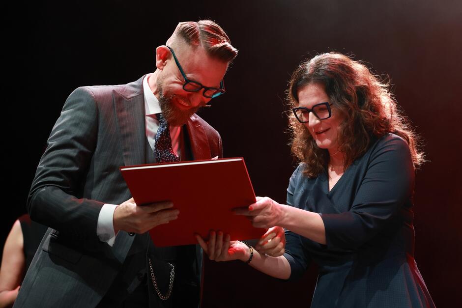roześmiany mężczyzna z brodą i w okularach przyjmuje z rąk kobiety dyplom