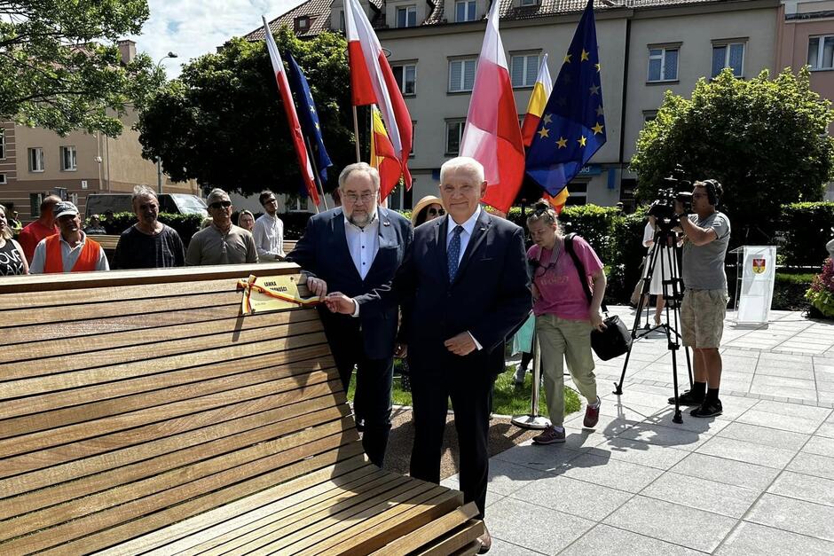 Dwaj mężczyźni przy dużej ławce, która jest po lewej. Na ławce jest kartka z nazwą skweru. W tle flagi Polski, Unii Europejskiej i Białegostoku