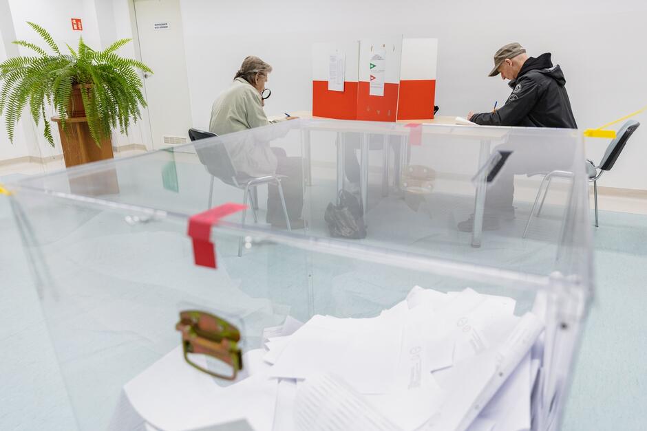 Zdjęcie przedstawia dwóch mężczyzn siedzących przy stole w lokalu wyborczym. Obaj są skupieni na wypełnianiu swoich kart do głosowania, które trzymają na stolikach z przegrodami zapewniającymi prywatność. W tle, po lewej stronie, znajduje się zielona paproć stojąca na drewnianym stojaku. Na pierwszym planie, nieco rozmyta, widoczna jest przezroczysta urna wyborcza, częściowo wypełniona kartami do głosowania.
