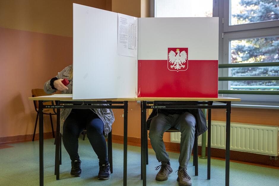 Zdjęcie przedstawia dwie osoby siedzące przy stole w lokalu wyborczym, zasłonięte parawanem wyborczym z godłem Polski i biało-czerwoną flagą. Osoby siedzą naprzeciwko siebie, po lewej stronie widać osobę w jasnej kurtce, która trzyma pieczątkę, a po prawej osobę w ciemnych ubraniach. Parawan dzieli ich na dwie oddzielne przestrzenie, zapewniając prywatność podczas głosowania. W tle znajduje się okno z widokiem na zewnętrzny krajobraz oraz grzejnik pod oknem. Ściany pomieszczenia mają pastelowe, jasnobrązowe kolory.