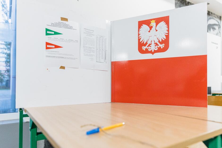 Zdjęcie przedstawia stanowisko do głosowania w Polsce. Na środku zdjęcia znajduje się drewniany blat z niebiesko-żółtym długopisem. Z prawej strony widoczna jest tablica w barwach flagi Polski, z godłem Polski przedstawiającym białego orła na czerwonym tle. Na lewej ścianie zamieszczone są dokumenty informacyjne, jeden z zielonym i czerwonym trójkątem oraz drugi zawierający tekst. W tle, przez okno, widać nieostre fragmenty budynku i drzewa. Cała scena jest dobrze oświetlona, sugerując, że zdjęcie zostało zrobione w ciągu dnia.