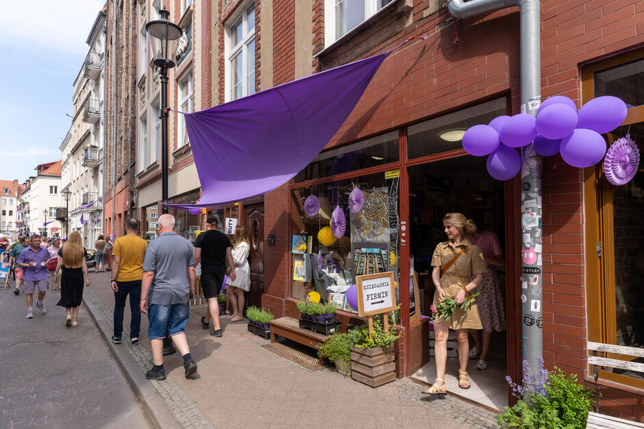 na zdjęciu fragment kilku kamienic w gdańsku, nad jedną z witryn sklepowych wiszą fioletowe balony i fioletowa firanka, wzdłuż ulicy widać kilku pieszych