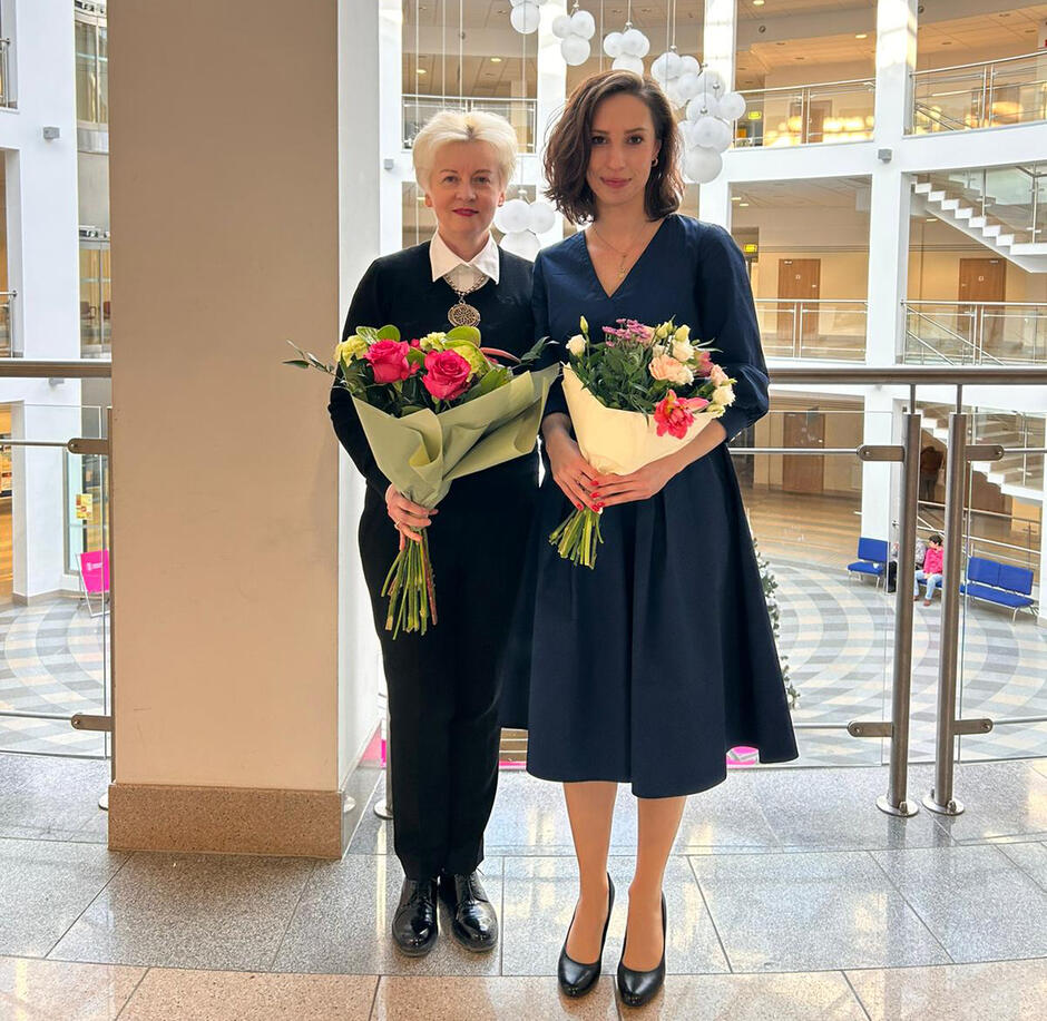 Dwie elegancko ubrane kobiety stoją w hallu uczelni z bukietami kwiatów w rękach. Jedna jest starsza, druga młodsza.