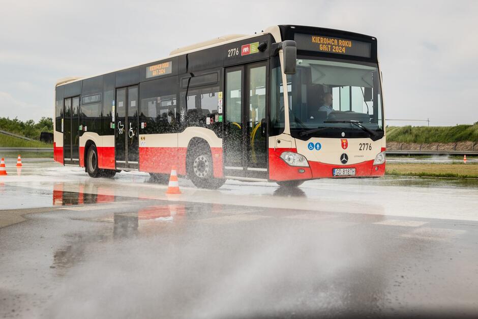 Autobus jedzie po torze z mokrą śliską nawierzchnią, spod kół pryskają krople wody. Na torze ustawione są pachołki. Przez szybę autobusu widać kierowcę. 
