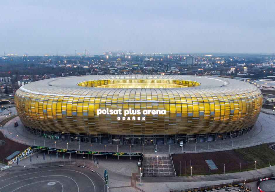 Na zdjęciu znajduje się stadion Polsat Plus Arena Gdańsk, o owalnym kształcie, z jasno podświetloną nazwą na elewacji. Otoczony jest miejską panoramą z rozproszonymi światłami w tle, co sugeruje, że zdjęcie zostało zrobione o zmierzchu.