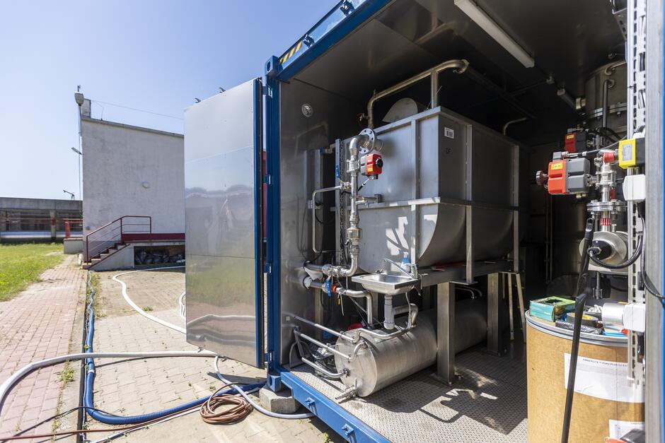 Na zdjęciu widoczny jest wnętrze kontenera technologicznego z projektu EMPEREST, który jest ustawiony na terenie przemysłowym, prawdopodobnie oczyszczalni ścieków. Wewnątrz kontenera znajdują się zaawansowane urządzenia do oczyszczania wody, w tym różne zbiorniki, rury i zawory. Kontener wyposażony jest w metalowe zbiorniki oraz system rur i pomp, które prawdopodobnie służą do procesów oczyszczania wody za pomocą technologii takich jak ozonowanie, adsorpcja na węglu aktywnym i wymiana jonowa. Widoczne są również urządzenia kontrolne i pomiarowe, które zapewniają prawidłowe funkcjonowanie systemu. Wnętrze kontenera jest dobrze oświetlone i wyposażone w różne akcesoria niezbędne do prowadzenia testów i operacji oczyszczania. Przed kontenerem znajdują się różne przewody i węże podłączone do systemu, wskazujące na aktywną pracę lub testy. W tle widoczna jest infrastruktura przemysłowa z betonowymi budynkami, schodami i rurami, co potwierdza lokalizację w obszarze przemysłowym związanym z oczyszczaniem ścieków.