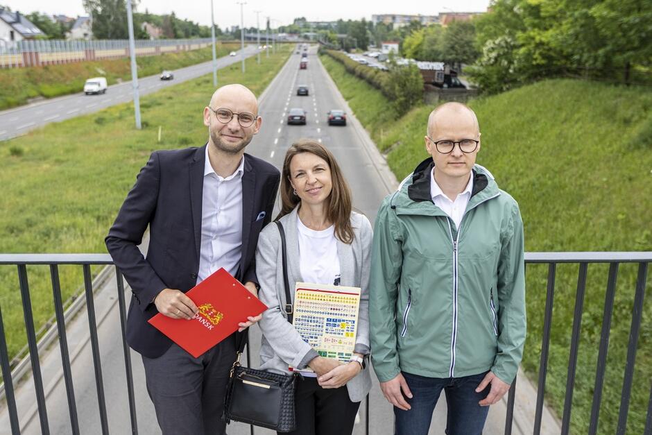Na zdjęciu widzimy trzy osoby stojące na moście nad dwupasmową drogą. Mężczyzna po lewej trzyma czerwoną teczkę z herbem Gdańska, kobieta w środku trzyma kolorową książkę, a mężczyzna po prawej ma na sobie zieloną kurtkę.
