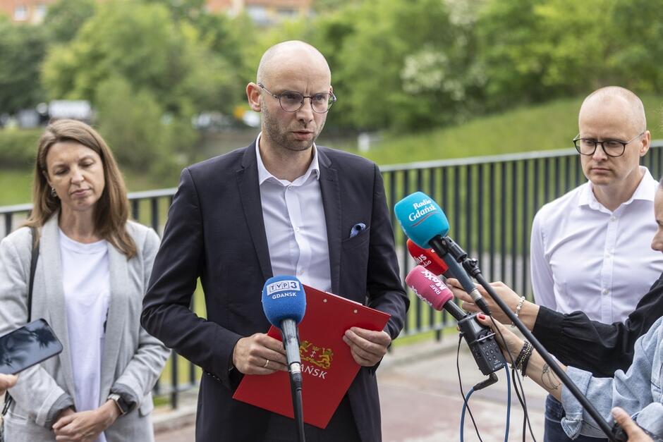 Na zdjęciu widzimy mężczyznę w garniturze, trzymającego czerwoną teczkę z herbem Gdańska, który udziela wywiadu mediom, przed mikrofonami oznaczonymi logotypami różnych stacji telewizyjnych i radiowych. Obok niego stoją kobieta i mężczyzna, słuchając wypowiedzi, a w tle widoczna jest zieleń.