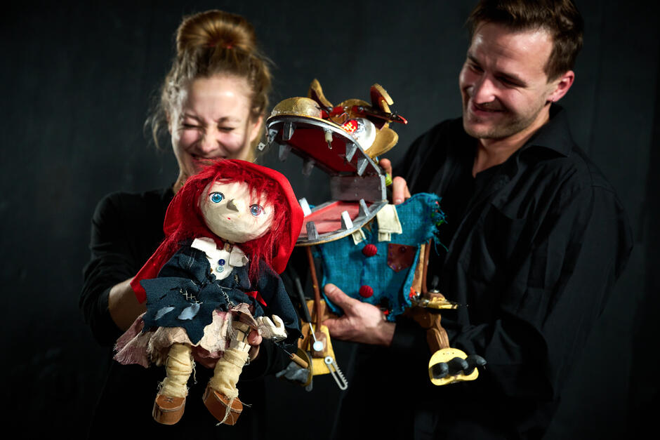 Zdjęcie przedstawia dwie osoby trzymające marionetki, jedną z nich jest lalka z czerwonymi włosami i dużymi, niebieskimi oczami, a drugą jest postać z otwartą paszczą, wyglądająca jak robotyczne zwierzę 